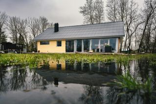 A picturesque rural house next to a reflecting pond surrounded by budding trees.